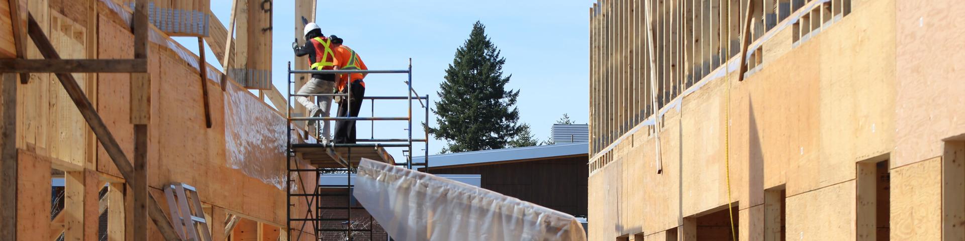 Two workers in safety gear stand atop a flat scaffolding working on a construction site.