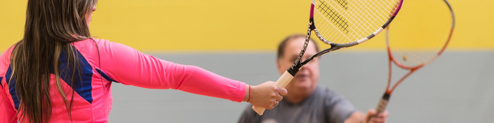 A woman in pink athletic wear holds out a badminton racket, while a man on the other side of the net matches her pose.