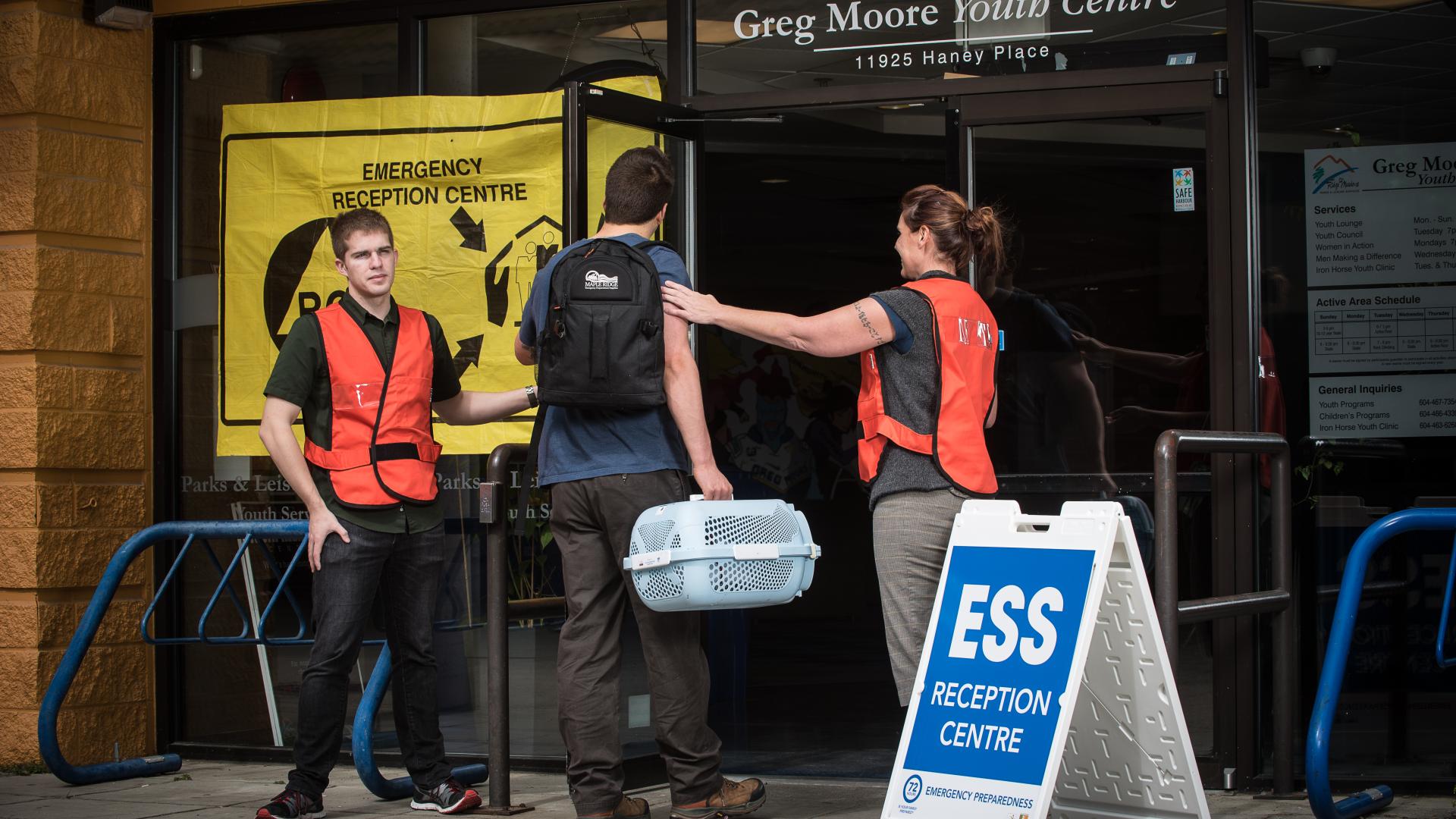 A pair of workers in high-visibility vests welcome a man carrying a small animal carrier to an Emergency Support Services reception centre.