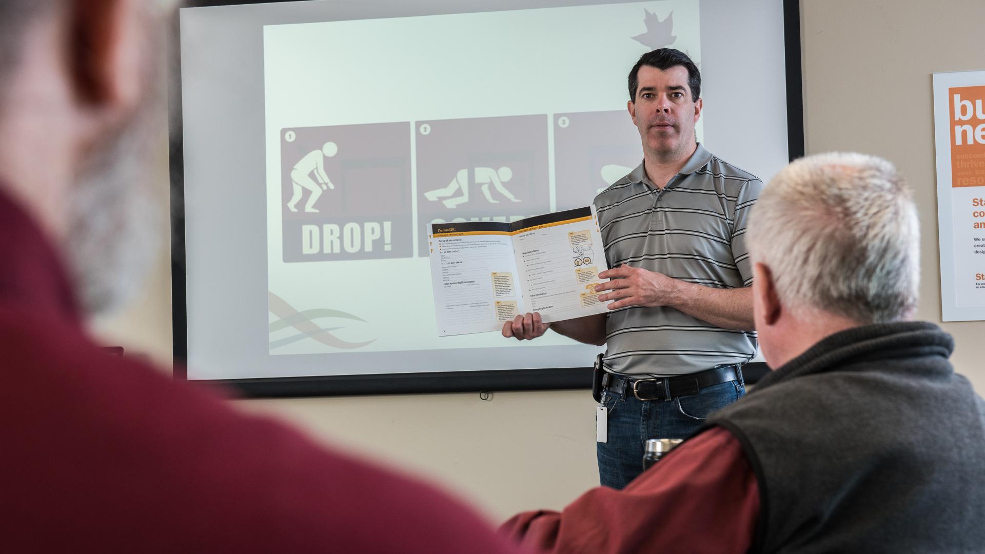 A man stands at the front of a classroom with a book open, and earthquake instructions on a screen behind him, teaching volunteers.
