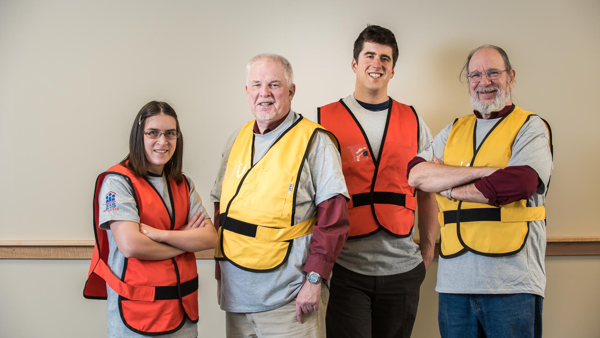 Four volunteers of varying ages, genders and heritages stand in alternating orange, yellow high-visibility vests.