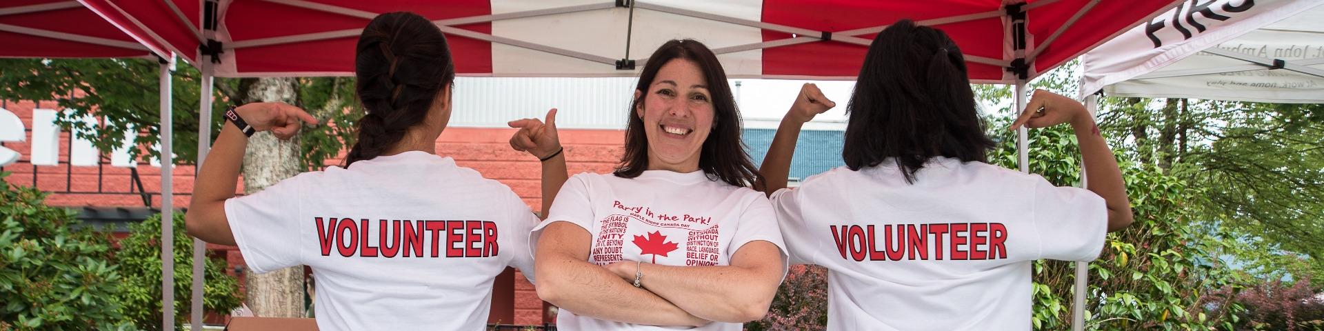 Two volunteers point to the word "volunteer" on the back of their shirts while a third smiles with crossed arms.