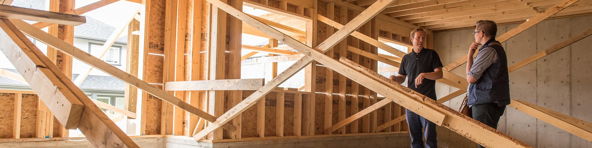 Two men in dark clothes stand inside a house under construction, having a discussion.