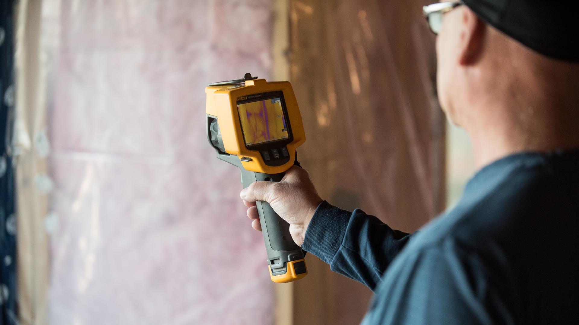 A man points a thermal imaging device at a wall of insulation.