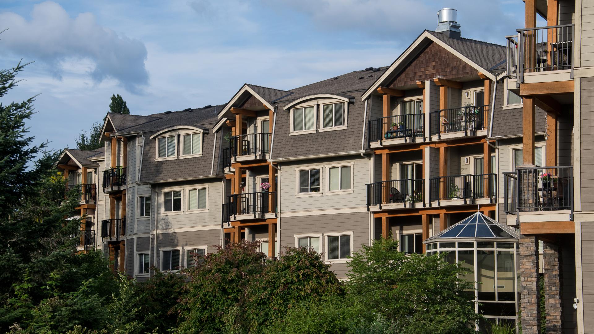 A grey apartment building with wooden accent sits behind a row of green trees.