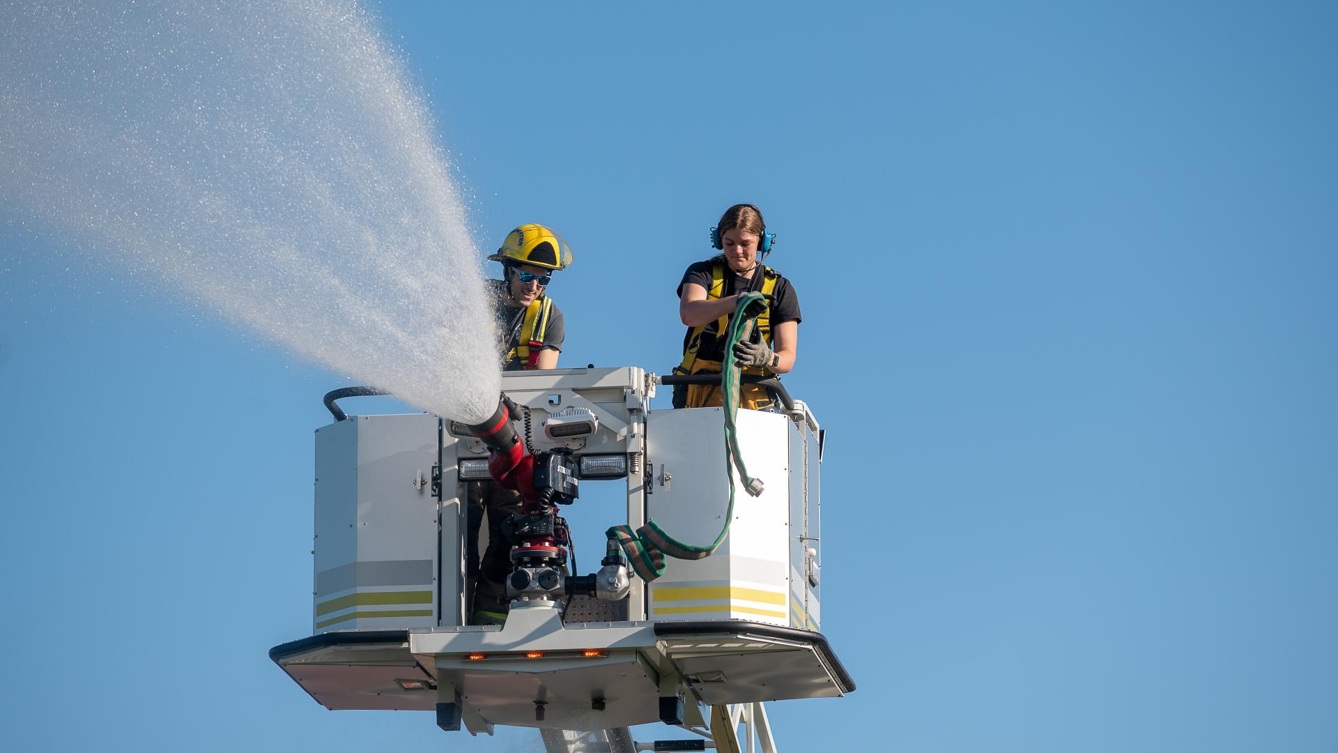 A pair of firefighters spray the hose from atop the truck's basket against a clear blue sky.