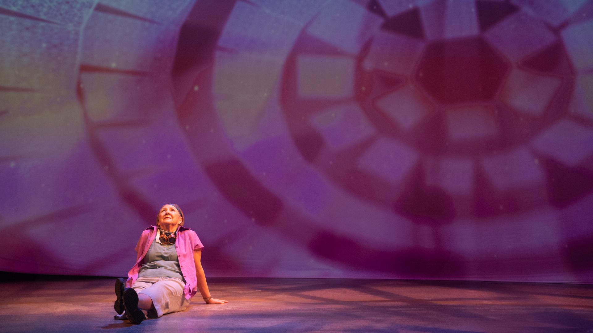 A female performer sits on a stage by herself under a kaleidoscopic set of lights.