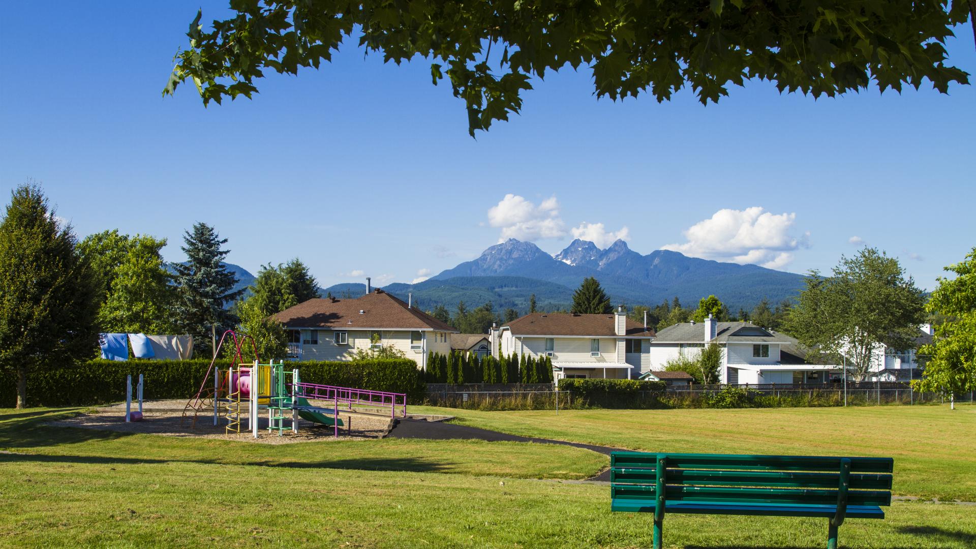 A small park and swing set sits downhill from a green park bench that rests in the shade of a tree.