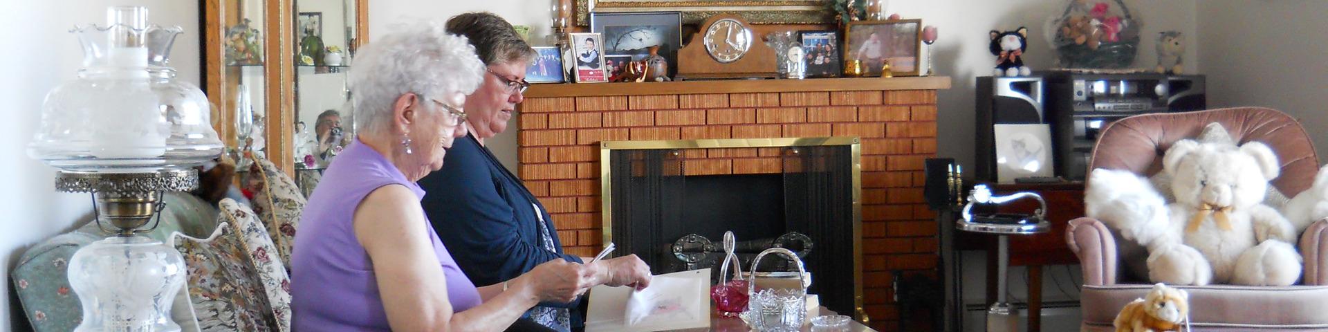 A senior woman sits on the couch with another woman, reviewing items in her home.