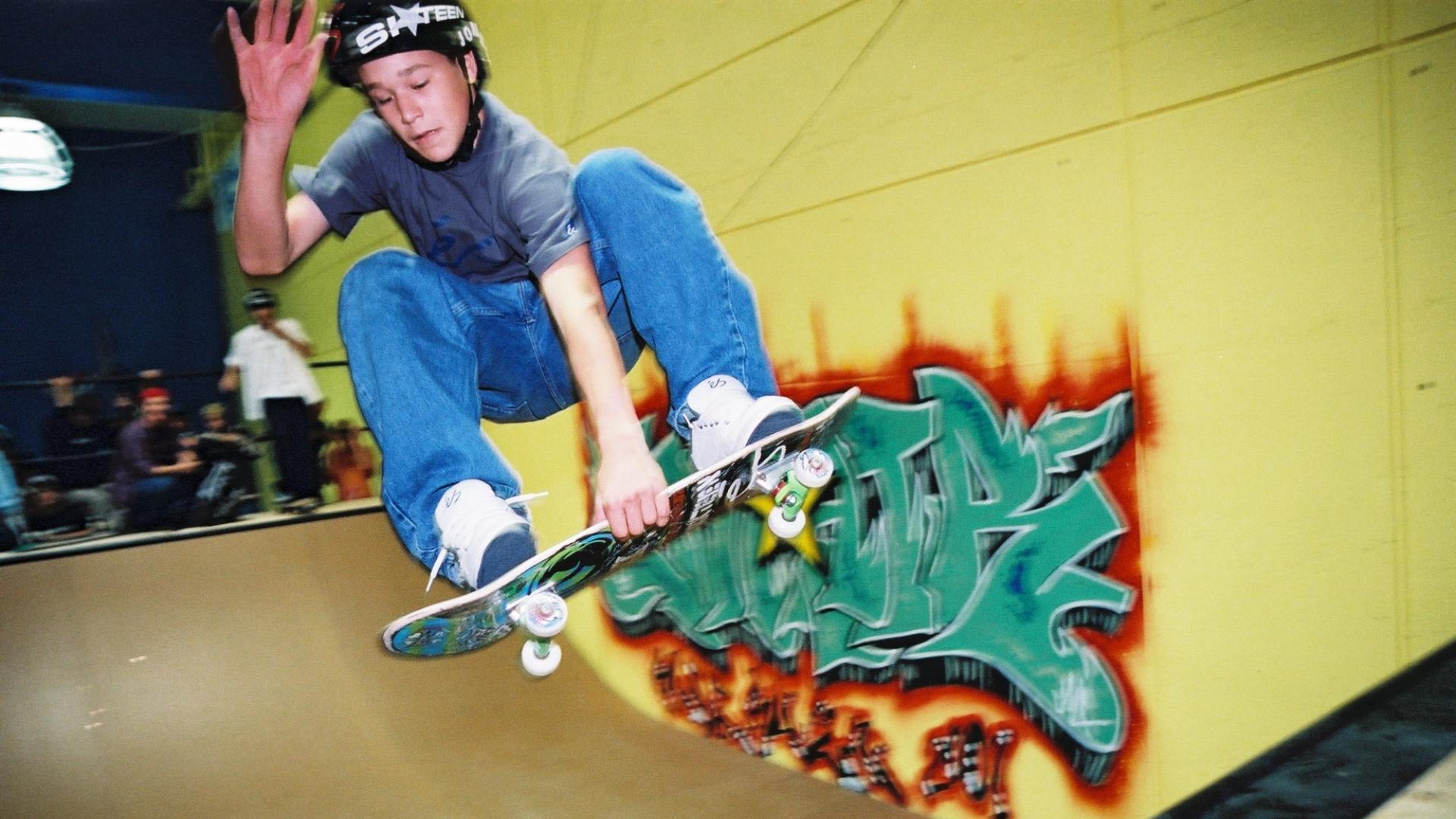 A boy on a skateboard with a black helmet soars through the air over a halfpipe. The wall is painted bright yellow with graffiti art on it, a 3 other boys stand at the top of the half pipe watching the first boy.