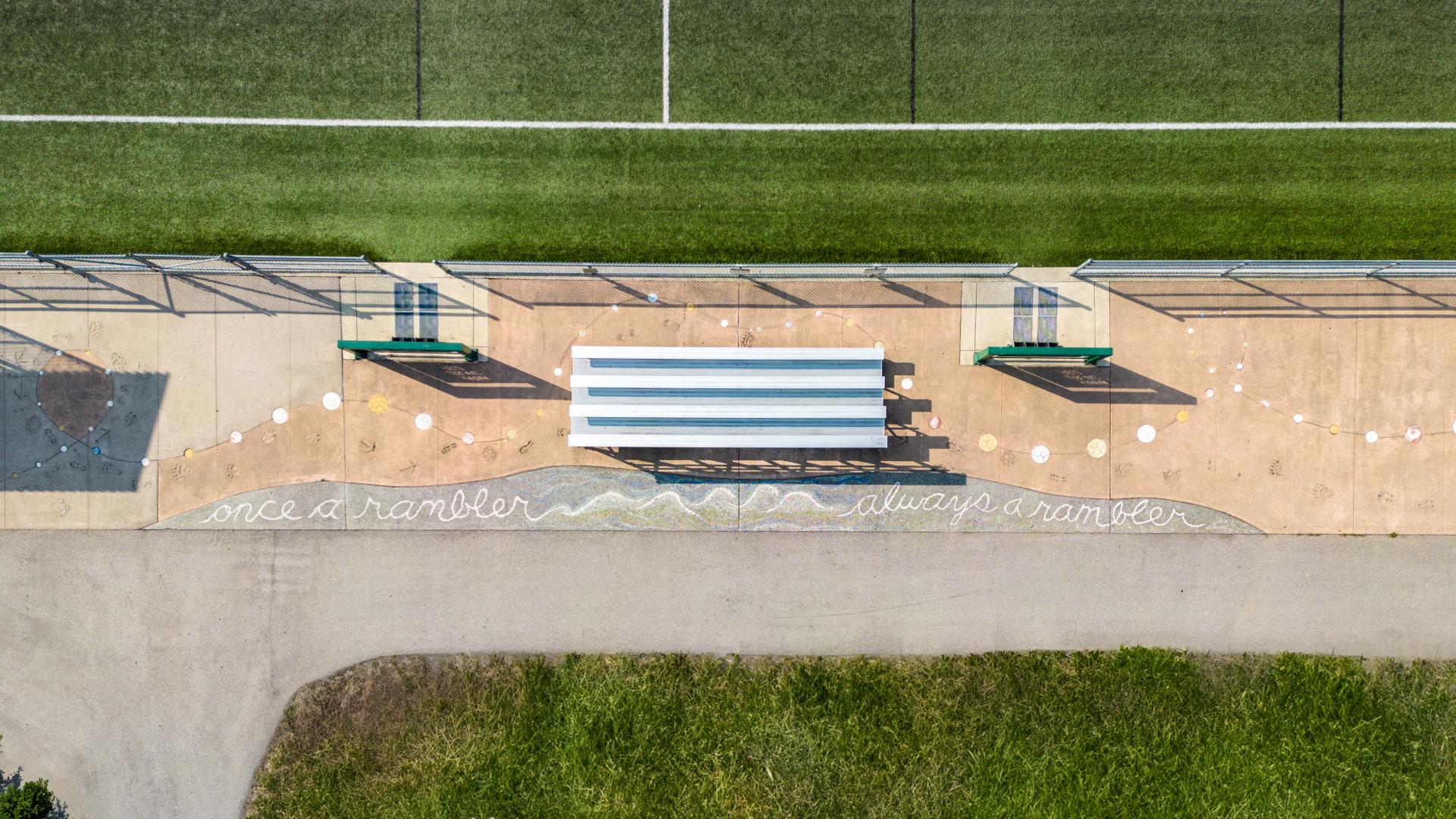 A bird's eye view of the public art installation at Karina LeBlanc Field in Merkley Park. A line of coloured soccer balls imprints are set into the concrete next to a paved pathway proclaiming "Once a rambler, always a rambler."