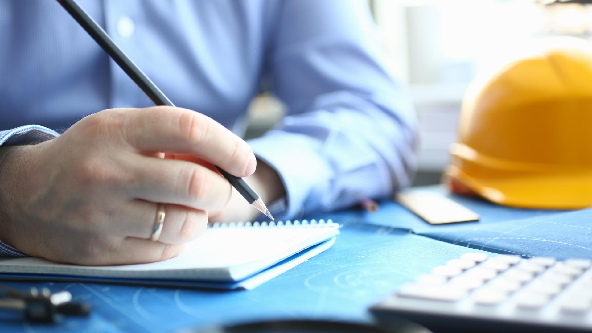 A married man makes notes on a pad of paper next to a calculator with a yellow hard hat in the background.