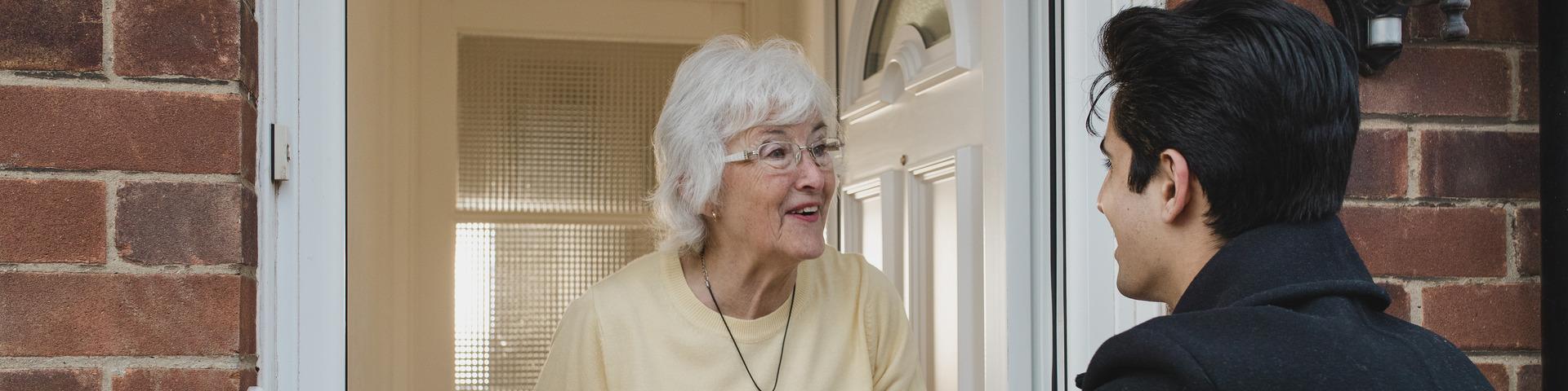 A senior woman greets a man at the door to her house.