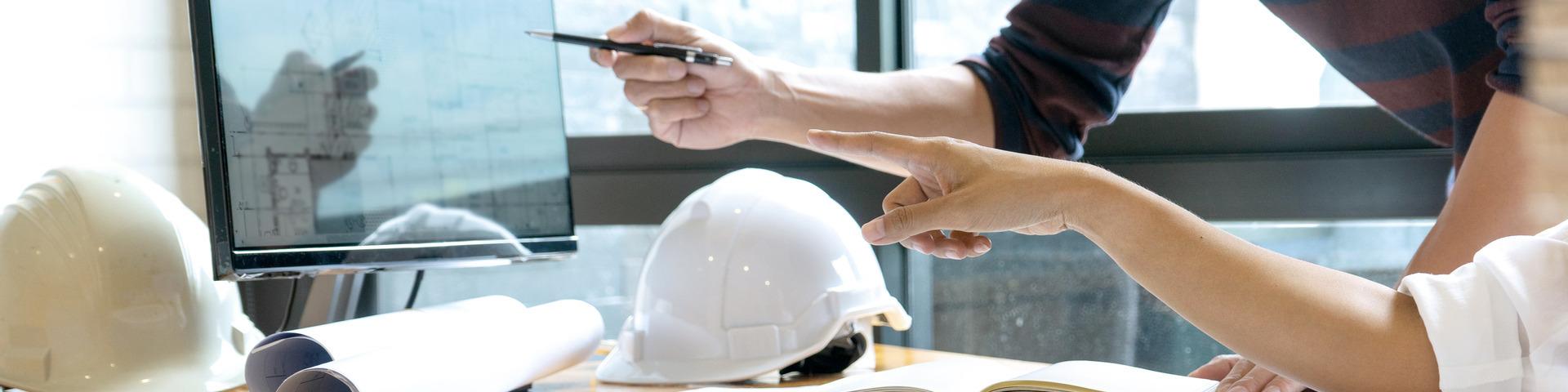 Two people point at plans on a computer screen, a white hard hat and physical paper plans on the table in front of them.
