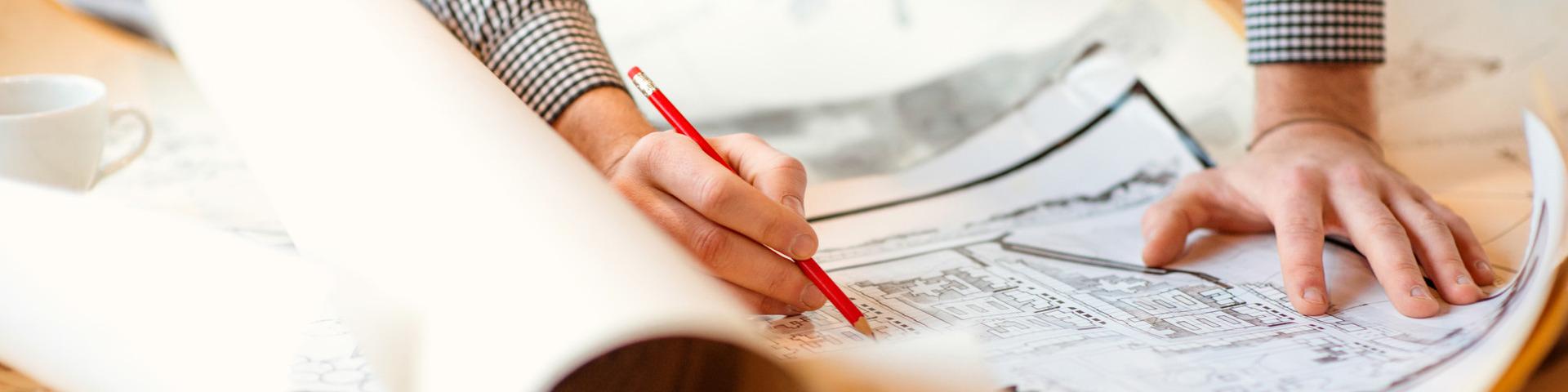 A person with a red pen spreads and marks now a note on a large paper plan.