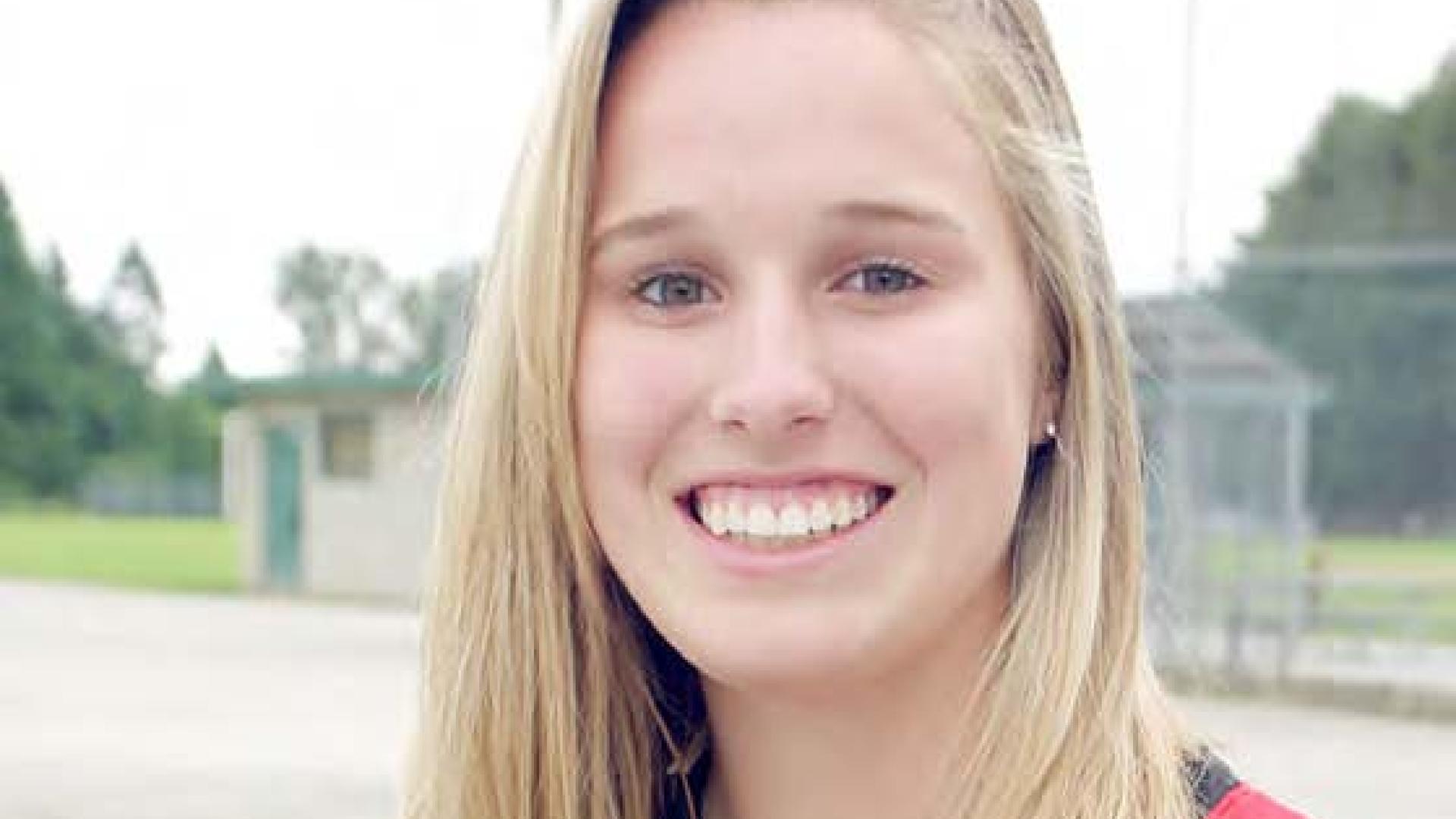Larissa Franklin smiles as she stands in an empty baseball diamond wearing a baseball glove.