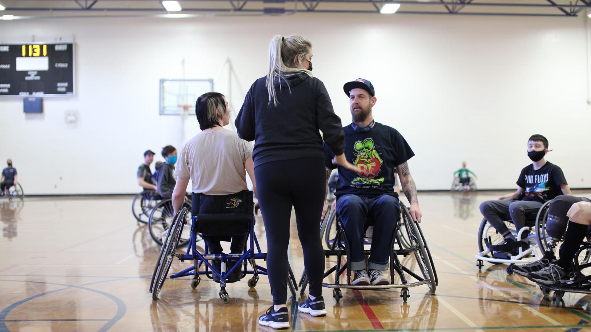 A referee facing two wheelchair basketball players on a gymnasium court