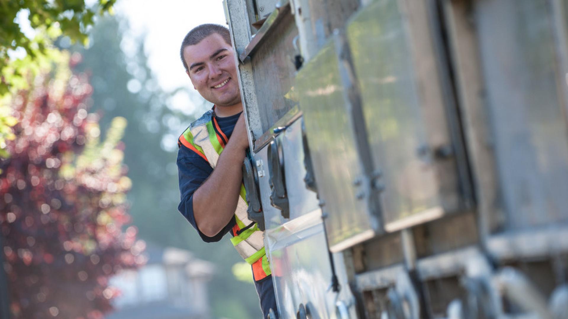 A curbside pick up recycling worker smiles as he holds onto the back of the recycling truck on his rounds.