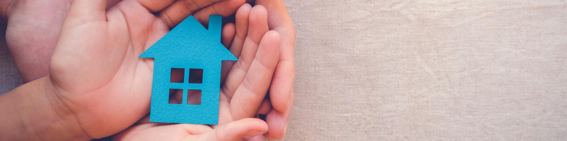 An adult cups their hands around a child's hands as they hold a blue stylized cut-out of a house.