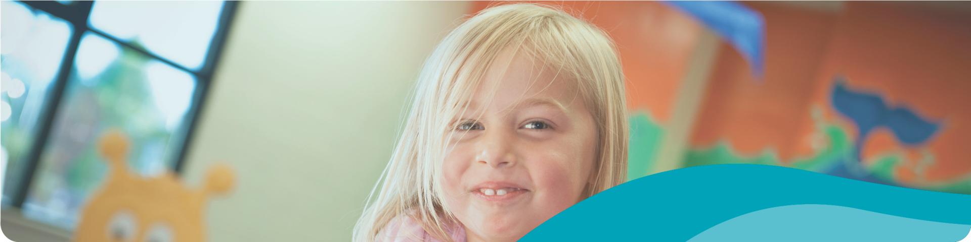 A young girl with blonde hair smiles at the camera. Behind her is a window and some wooden cabinets.
